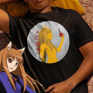 以下が含まれることがあります： 黒いTシャツ。円形のグラフィックには、赤いリンゴを持った黄色いアニメスタイルのキャラクターが描かれています。グラフィックには日本語のテキストと水色の背景が含まれています。キャラクターはオオカミの耳を持っています。カジュアルウェアアイテムです。