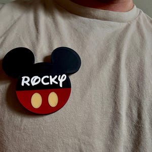May include: A black, red, and white Mickey Mouse-shaped name tag with the name "Rocky" in white lettering. The name tag has two yellow ovals representing Mickey's nose. The name tag is attached to a beige t-shirt.