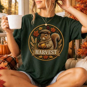 May include: A dark green t-shirt featuring a circular graphic of a squirrel wearing a cap and sweater, holding an acorn. The design includes autumn leaves, wheat stalks, and a banner with the text HARVEST. The shirt is worn with a gold necklace.