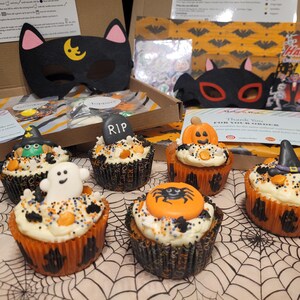 May include: A Halloween cupcake decorating kit featuring six cupcakes topped with white frosting, sprinkles, and edible figures including a ghost, a tombstone with RIP, a pumpkin, a spider, and witch hats, alongside two cat masks and instruction cards.
