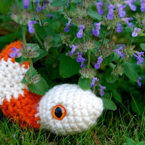 Op de afbeelding: Een gehaakte oranje en witte slang met een groot oranje oog, liggend in een bed van groen gras en paarse bloemen.