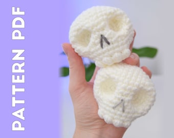 May include: Two white crocheted skull ornaments with black eyes. The skulls are held in a hand with a green plant in the background.