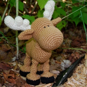 May include: A handmade crochet moose toy. The moose is primarily brown with white antlers and black hooves. It is standing in a natural outdoor setting with green leaves and brown foliage in the background.