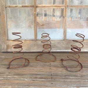 May include: Three rusty metal springs, each with a coiled top and a circular base. The springs are arranged on a weathered wooden surface, with a window in the background.