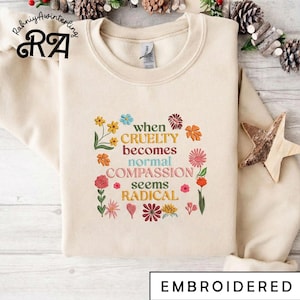 Op de afbeelding: Crèmekleurige geborduurde sweatshirt met de tekst "when cruelty becomes normal compassion seems radical" omringd door kleurrijke bloemmotieven. Het woord "EMBROIDERED" staat onderaan.