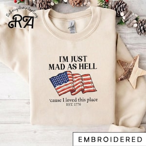 May include: Cream-colored sweatshirt with the embroidered text "I'M JUST MAD AS HELL" and an American flag graphic. Below the graphic is the text "'cause I loved this place EST. 1776". The word "EMBROIDERED" is at the bottom.