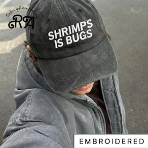 Puede incluir: Gorra de béisbol gris oscuro bordada con el texto "SHRIMPS IS BUGS" en blanco. La gorra tiene una visera curva y está hecha de una tela desgastada y texturizada. La palabra "EMBROIDERED" está en la parte inferior de la imagen.