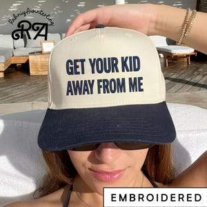 May include: A cream-colored baseball cap with a navy blue brim. The cap features the embroidered text "GET YOUR KID AWAY FROM ME" in navy blue block letters. The word "EMBROIDERED" is also visible.
