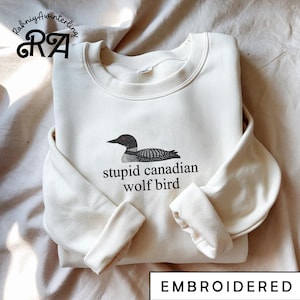 Pode incluir: Camisola bordada creme com um gráfico de pássaro a preto e branco e o texto "stupid canadian wolf bird". A camisola tem gola redonda e mangas compridas. A palavra "EMBROIDERED" está na parte inferior.