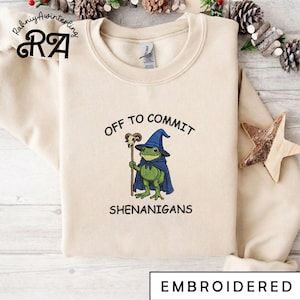 Può includere: Felpa color crema con un design ricamato raffigurante una rana verde con cappello e mantello da mago blu, che tiene un bastone. Il testo recita "OFF TO COMMIT SHENANIGANS". La parola "RICAMATO" è in basso.