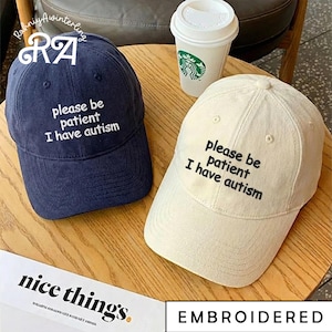 May include: Two baseball caps, one navy blue and one off-white, are displayed. Both caps have the embroidered text "please be patient I have autism". A white paper coffee cup with a green logo is in the background. The caps are on a wooden surface.