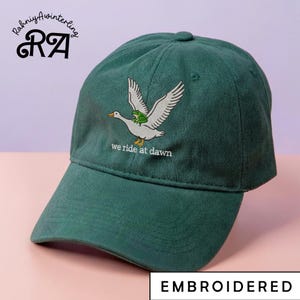 Pode incluir: Boné de beisebol verde escuro com um desenho bordado de um ganso branco carregando um sapo verde, com o texto "we ride at dawn". O boné também apresenta o texto "EMBROIDERED" e o logotipo "RA".