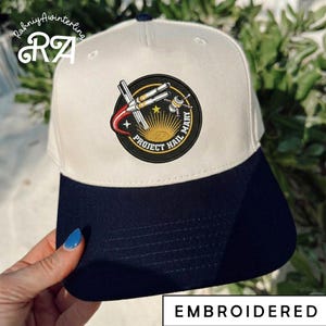 May include: A white and navy blue baseball cap with an embroidered patch featuring a rocket, satellite, and the text "PROJECT HAIL MARY". The cap has the word "EMBROIDERED" at the bottom.