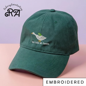 May include: A dark green baseball cap with an embroidered design of a white duck with a green frog on its back. The text "we ride at dawn" is below the duck. The word "EMBROIDERED" is at the bottom.