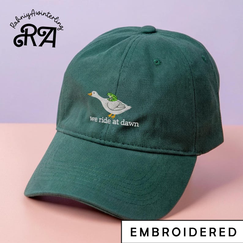 May include: A dark green baseball cap with an embroidered design of a white duck with a green frog on its back. The text "we ride at dawn" is below the duck. The word "EMBROIDERED" is at the bottom.