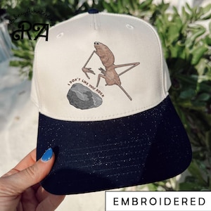May include: A white and navy blue baseball cap with an embroidered cartoon creature and the text "I don't like this rock." The cap has a speckled design on the brim and the word "EMBROIDERED" is printed on the bottom.