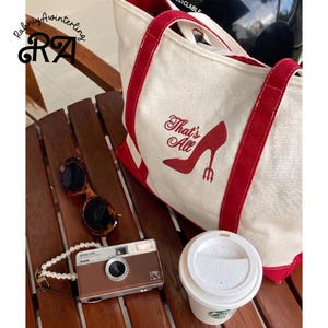 May include: A cream tote bag with red handles and trim, printed with "That's All" and a high-heeled shoe. Also, a vintage brown Kodak camera, sunglasses, a pearl necklace, and a coffee cup.