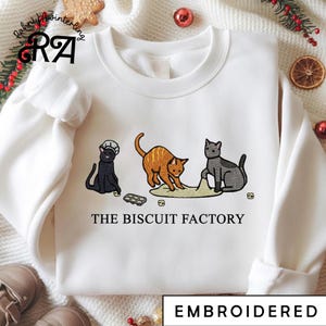 May include: White sweatshirt with an embroidered design featuring three cats. The cats are in black, orange, and gray, with the text "THE BISCUIT FACTORY" below. The word "EMBROIDERED" is at the bottom.