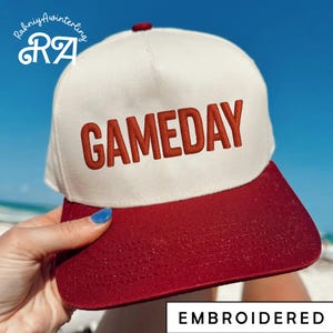 May include: A white and red baseball cap with the word "GAMEDAY" embroidered in red. The cap has a red brim and a white crown. The word "EMBROIDERED" is written in a white rectangle at the bottom of the image.