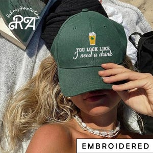 以下が含まれることがあります： 「YOU LOOK LIKE I need a drink」の刺繍とビールのグラフィックが入った、濃い緑色の野球帽。つばはカーブしており、上部にボタンが付いています。「EMBROIDERED」の文字が下部にあります。