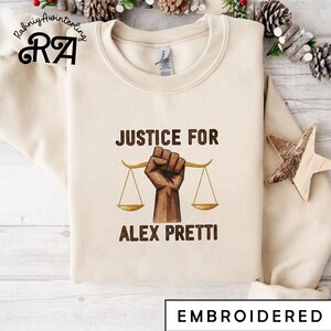May include: A cream-coloured sweatshirt featuring the embroidered text "JUSTICE FOR ALEX PRETTI" above a brown fist holding scales. The word "EMBROIDERED" is at the bottom. The sweatshirt has a crew neck.