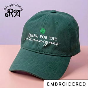 May include: A dark green baseball cap with a curved peak. The cap has the embroidered text "HERE FOR THE shenanigans" in white, and a green shamrock. The word "EMBROIDERED" is at the bottom.