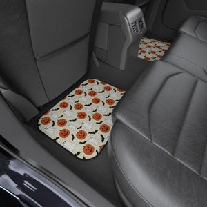 May include: A set of rectangular car floor mats featuring a repeating pattern of orange jack-o'-lantern pumpkins, white ghosts, and black bats on a light cream-colored background. The mats are positioned on the floor of a vehicle interior.