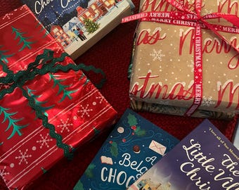 May include: A collection of wrapped gifts with a Christmas theme. The gifts are wrapped in red, brown, and purple paper with festive ribbons and snowflakes. The books are titled "Starry Skies Over The Chocolate Pot Cafe", "A Belgian Chocolate Christmas", and "The Little Village Christmas".