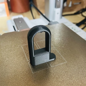 May include: A black plastic 3D printed hook with a rounded rectangular base. The hook has a smooth, matte finish.