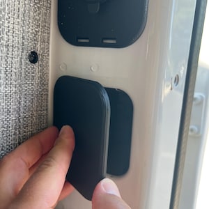 May include: A black rectangular cover is being opened, revealing a recessed area with a USB port and a blue light. The cover is being opened by a hand. The cover is mounted on a white surface.