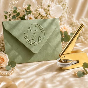 May include: A sage green envelope featuring a circular embossed monogram with the letters A and B, an ampersand, and the date 26.10.2025, accompanied by a gold-toned metal embosser tool positioned to the right of the envelope.