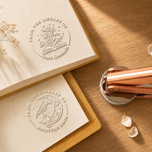 May include: A copper-colored metal embosser tool next to two books with embossed circular designs. One design features flowers emerging from an open book with the text FROM THE LIBRARY OF Sami Germain. The other shows a raven, moon, and skull with FROM THE LIBRARY OF JONATHAN COOK.