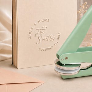 May include: A light mint green metal embosser tool positioned next to a beige cardstock paper. The paper features a circular embossed design with the text James & Mason, The Smiths, and the date November 12, 2025, pressed into the surface.