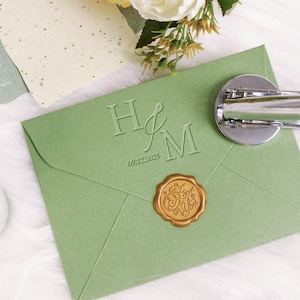 May include: A sage green wedding invitation envelope with embossed initials "H&M" and the date "09.12.2025". A gold wax seal with script text is affixed. A silver wax seal stamp is on the envelope. Decorative paper and flowers are in the background.
