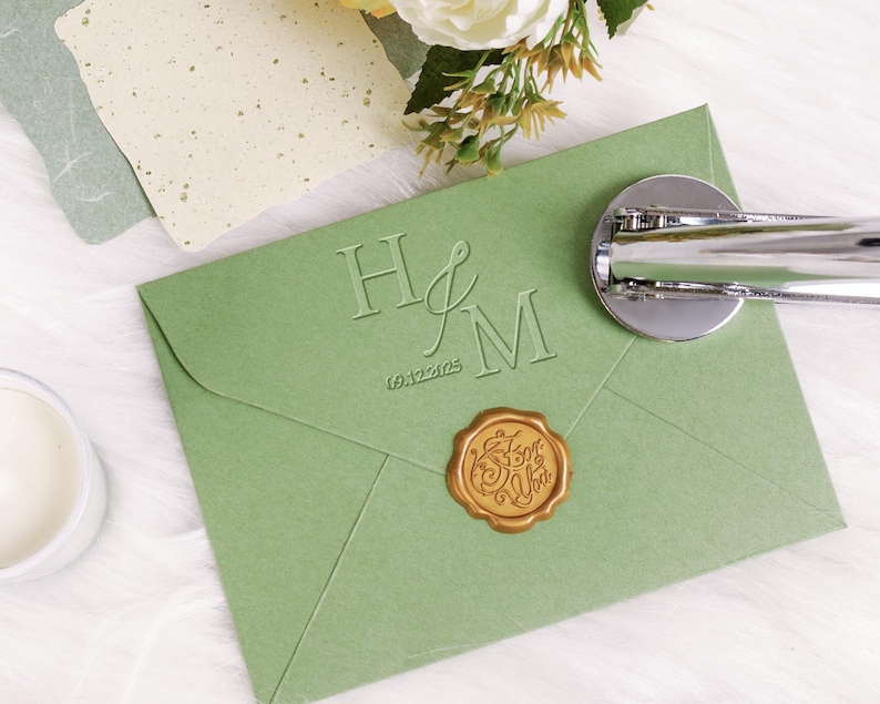 May include: A sage green wedding invitation envelope with embossed initials "H&M" and the date "09.12.2025". A gold wax seal with script text is affixed. A silver wax seal stamp is on the envelope. Decorative paper and flowers are in the background.
