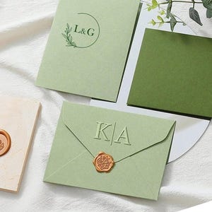 May include: Light green wedding invitation suite with embossed initials and a wax seal. The suite includes envelopes, cards, and a decorative wax seal. The design features a minimalist aesthetic with a focus on elegant typography and a natural color palette.