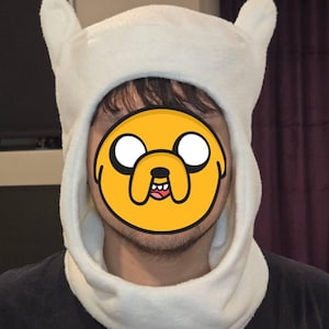 May include: A white plush hat with two upright ears and a neck warmer. The hat is worn by a person, with a yellow cartoon face covering the face. The hat is made of a soft, fuzzy material.