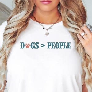 May include: White t-shirt featuring the text "DOGS > PEOPLE" in teal with a red paw print. The casual tee is suitable for everyday wear. The design is centrally placed on the front of the shirt.