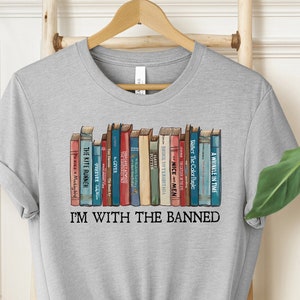 May include: Gray t-shirt with a graphic of a bookshelf full of books. The text "I'm with the banned" is printed below the bookshelf.