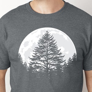 Peut inclure: Un t-shirt gris foncé chiné avec une silhouette blanche d'un pin devant une pleine lune. L'arbre est entouré de petits arbres.