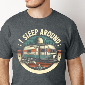 May include: A dark grey t-shirt with a vintage-style graphic of a camper van and the text "I Sleep Around". The graphic features a full moon, stars, and trees.