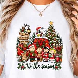 May include: A white t-shirt featuring a colourful graphic of Peanuts characters in a Christmas setting with a decorated tree, a red sleigh, and signs reading Hot Cocoa, Sleigh Rides, and Tree Farm, with the text 'tis the season below.