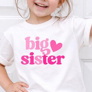 May include: A smiling young girl wears a white t-shirt with the words "big sister" in pink and a pink heart. She is wearing blue denim shorts and a pink bow in her hair.