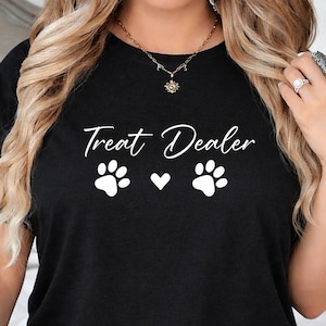 May include: Black t-shirt featuring the text "Treat Dealer" in a cursive font, accompanied by two white paw prints and a heart symbol. The top has a crew neck.