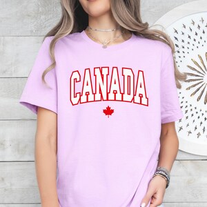 Peut inclure: Un t-shirt violet clair avec le mot "CANADA" en lettres blanches avec un contour rouge. Une feuille d'&eacute;rable rouge se trouve sous le mot.