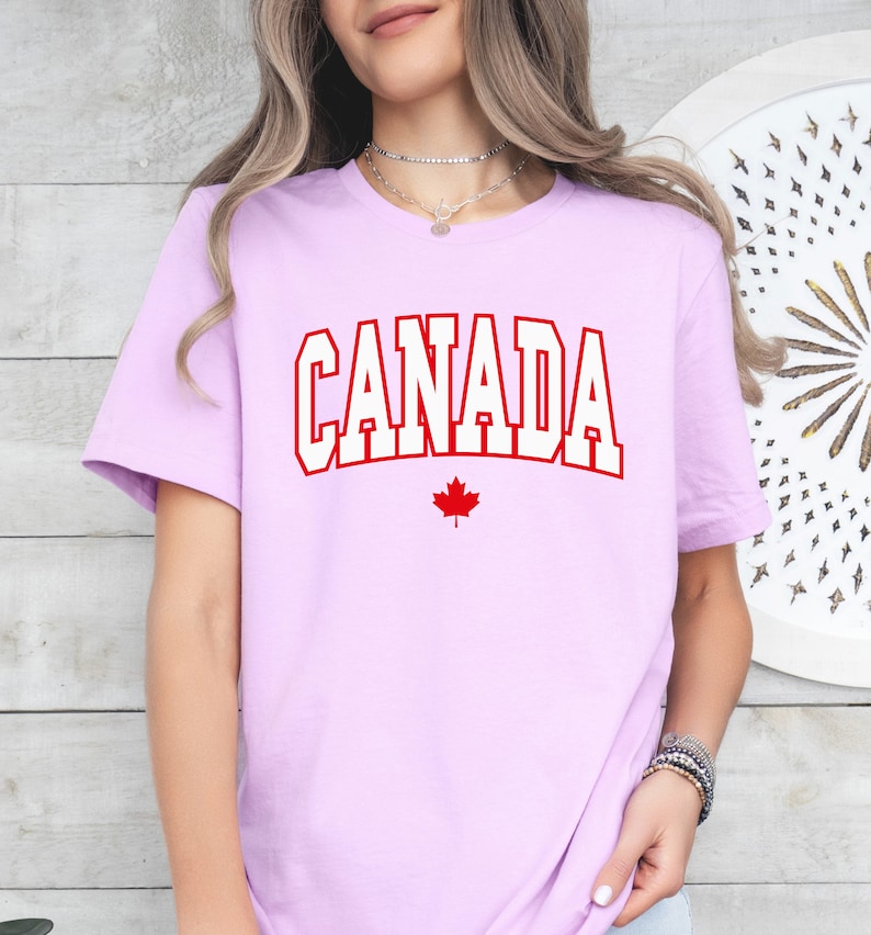 Peut inclure: Un t-shirt violet clair avec le mot "CANADA" en lettres blanches avec un contour rouge. Une feuille d'&eacute;rable rouge se trouve sous le mot.