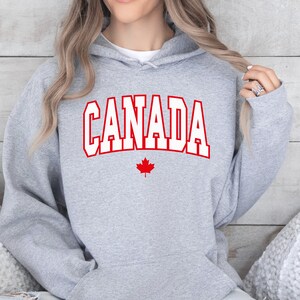 Peut inclure: Sweat-shirt &agrave; capuche gris avec le mot "CANADA" en lettres blanches sur fond rouge et une feuille d'&eacute;rable rouge en dessous.