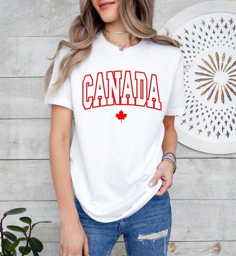 Peut inclure: T-shirt blanc avec le texte rouge "CANADA" et une feuille d'&eacute;rable rouge en dessous.
