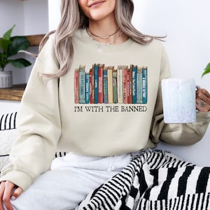 May include: A beige sweatshirt with a graphic of a bookshelf full of books. The text "I'm with the banned" is printed below the bookshelf graphic.
