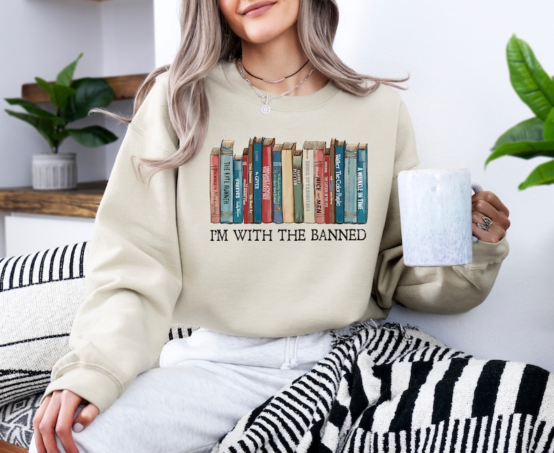 May include: A beige sweatshirt with a graphic of a bookshelf full of books. The text "I'm with the banned" is printed below the bookshelf graphic.